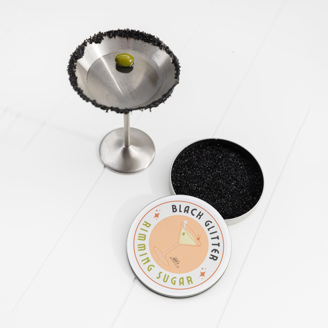 A silver martini glass with black sugar on the rim next to an open container of black glitter rimming sugar.