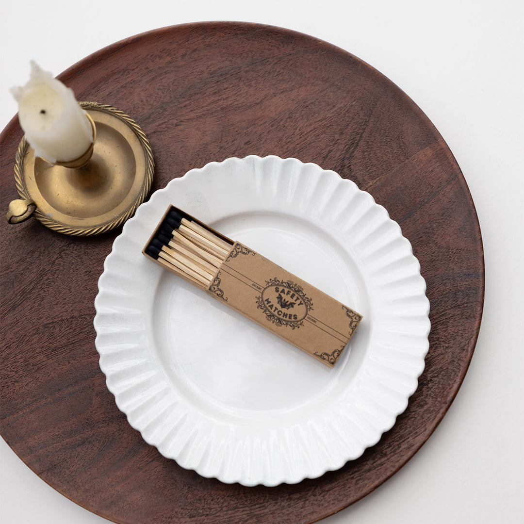 An open box of safety matches lays on a white circular plate on a dark wooden platter next to a candle.