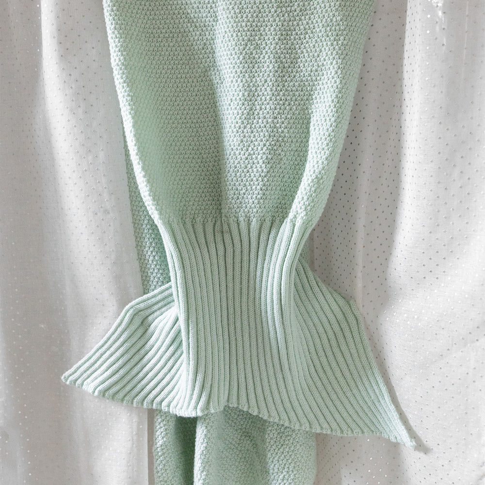 a seafoam green, knit mermaid tail blanket lays on a white background