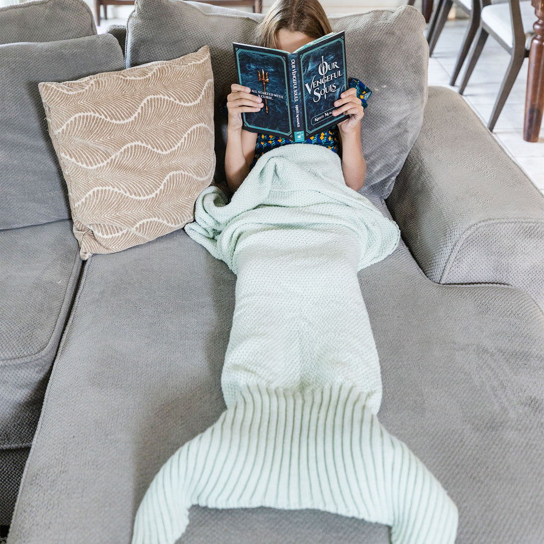 a white girl lays on a gray couch in a seafoam green, knit mermaid tail blanket reading a hardcover copy of Our Vengeful Souls