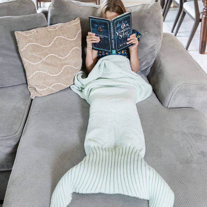a white girl lays on a gray couch in a seafoam green, knit mermaid tail blanket reading a hardcover copy of Our Vengeful Souls
