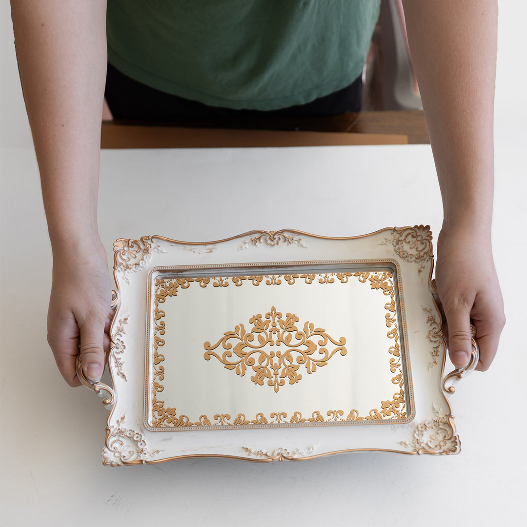 Hands holds out a white mirrored tray with gold accents over a white table.