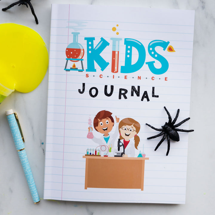 a notebook labeled Kids Science Journal with a cartoon image on the front of two children at a desk with science equipment. On top of the notebook is a fake black spider and next to the notebook is a pen