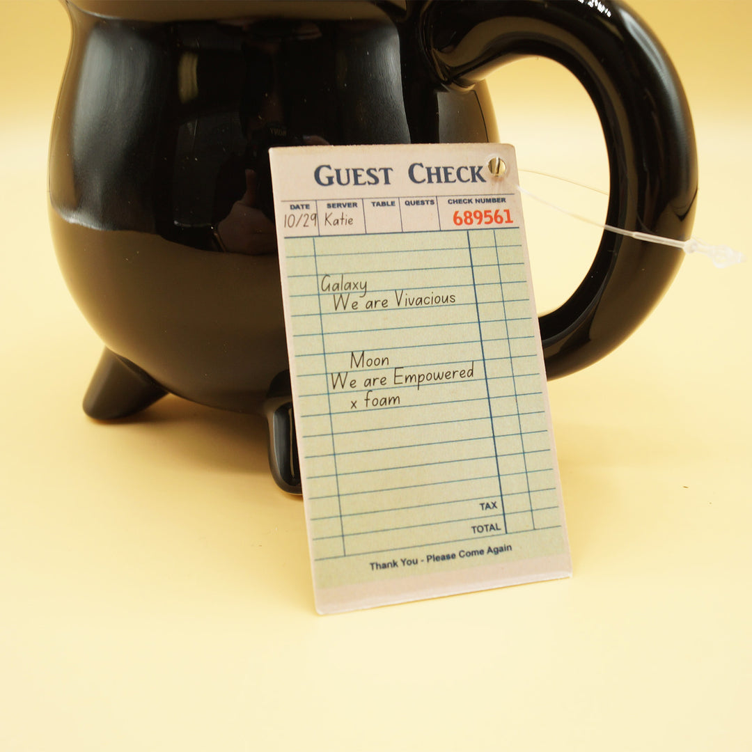 a small piece of paper labeled Guest Check with check-in information on it sits against a black cauldron-shaped mug