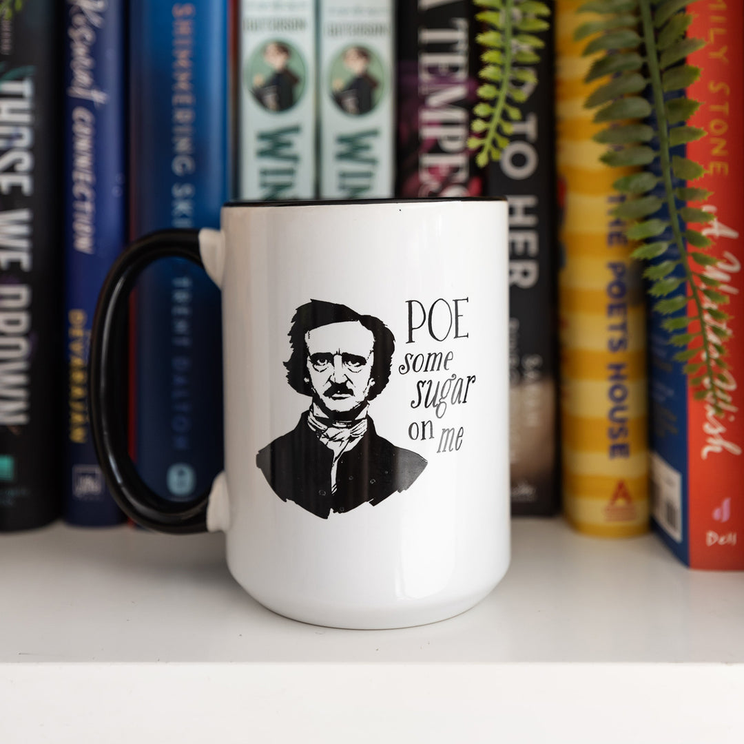 A white mug with a black handle sits on a bookshelf. On the front of the mug is a drawing of Edgar Allen Poe and the quote POE some sugar on me
