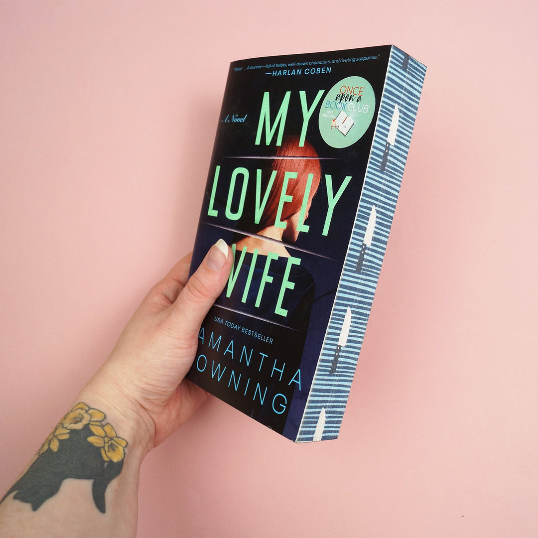 Book titled 'My Lovely Wife' by Samantha Downing showcasing patterned page edges is held by a hand against a pink background