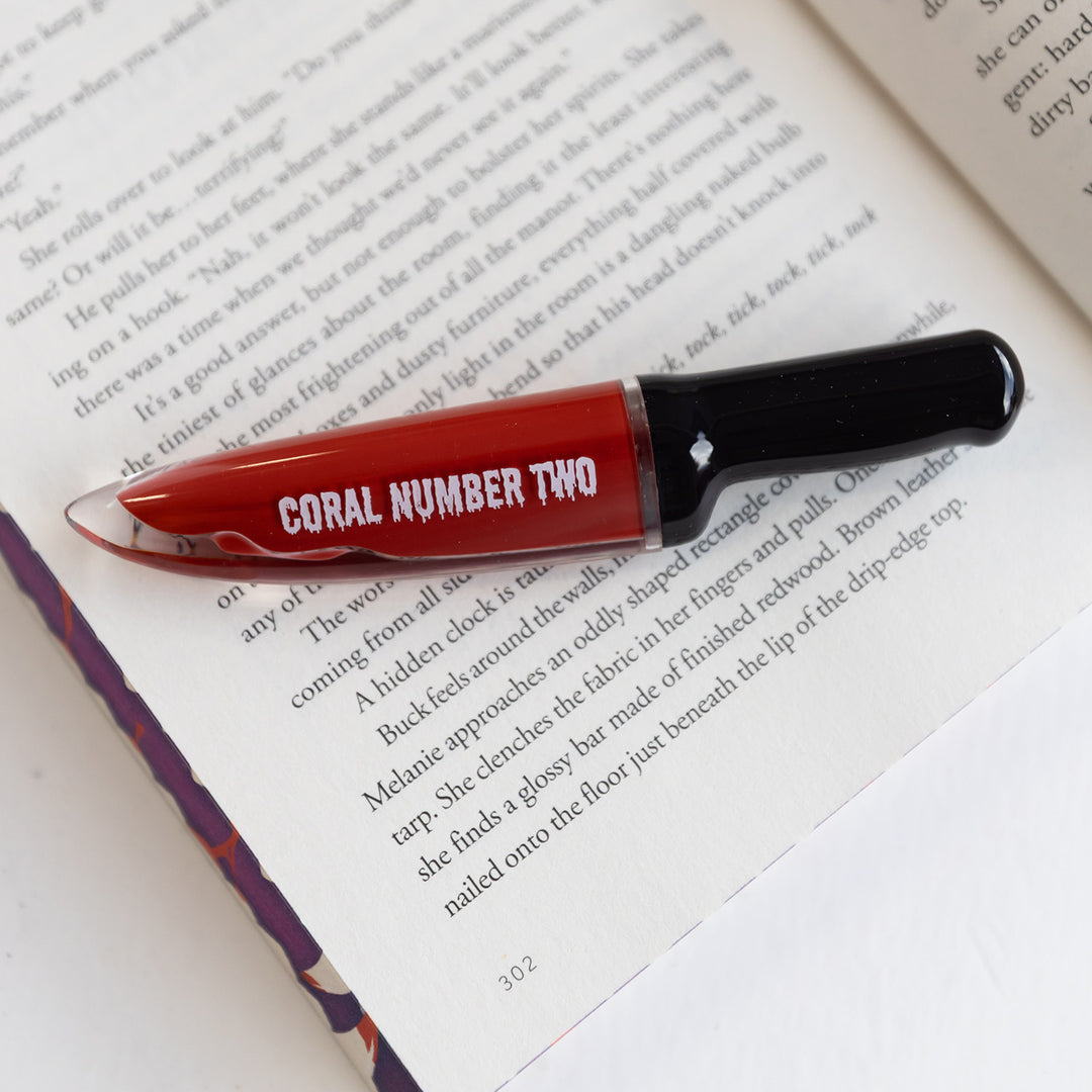 A knife-shaped bottle of red lipstick that says Coral Number Two on the side lays on an open book.