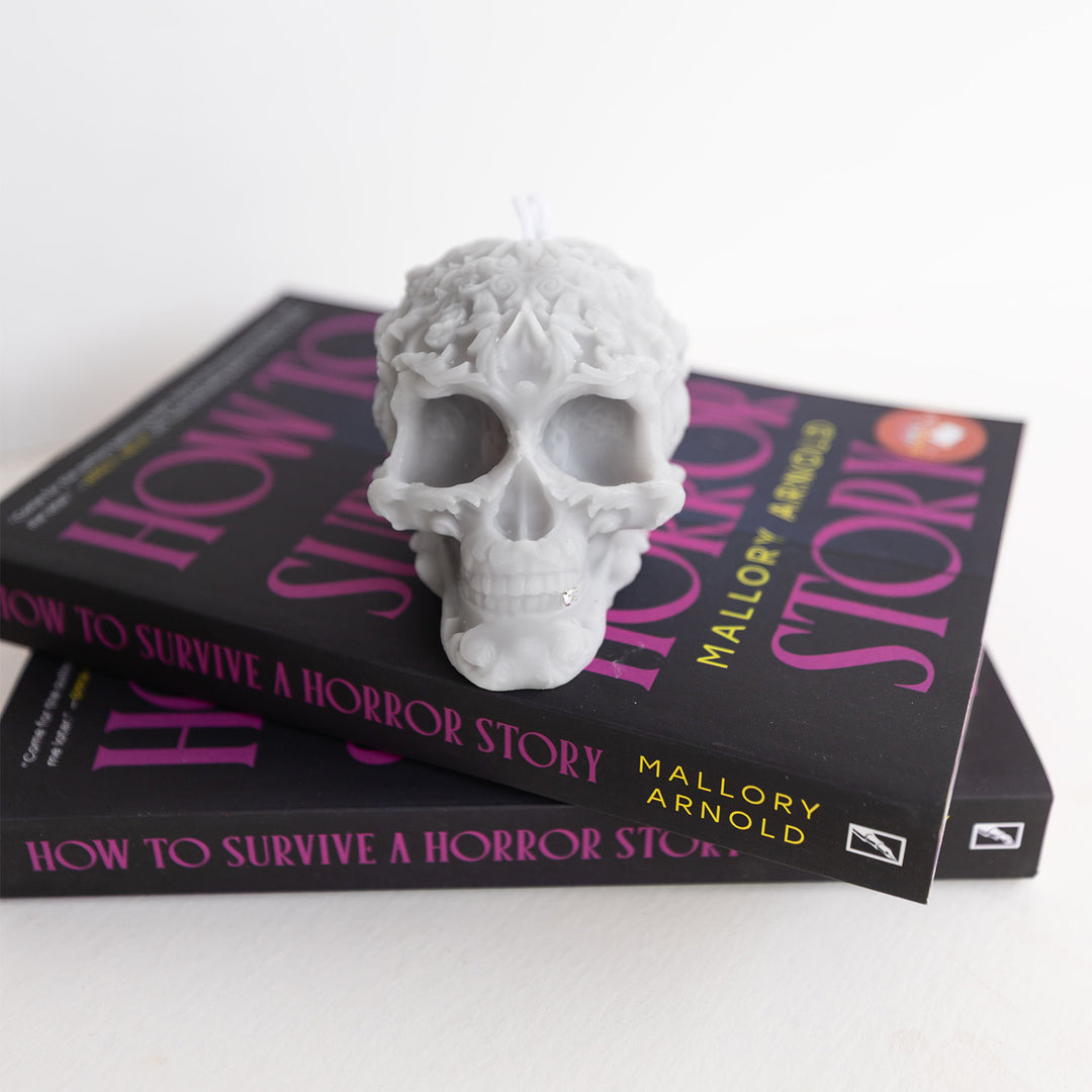 A gray filigree-patterned skull candle with a silver tooth sits on a stack of two paperback editions of How to Survive a Horror Story.