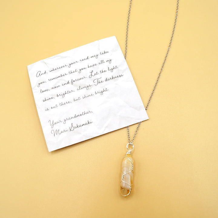 A yellow and clear crystal on silver chain next to a card that reads "And, wherever your road may take you, remember that you have all my love, now and forever. Let the light shine, brighter, always. The darkness is out there, but shine bright. Your grandmother, Mari Sakamaki.