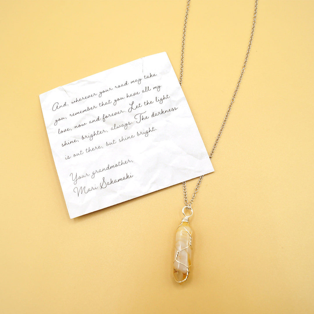 A yellow and clear crystal on silver chain next to a card that reads "And, wherever your road may take you, remember that you have all my love, now and forever. Let the light shine, brighter, always. The darkness is out there, but shine bright. Your grandmother, Mari Sakamaki.