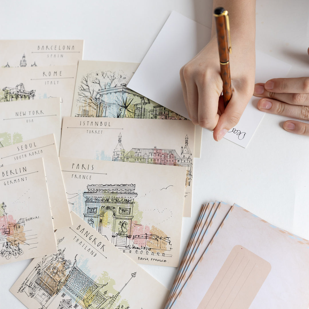 A hand writes on the back of a postcard with a pen. Next to the postcard are a set of nine more postcards with an illustration of a different city on each with a stack of envelopes next to them.