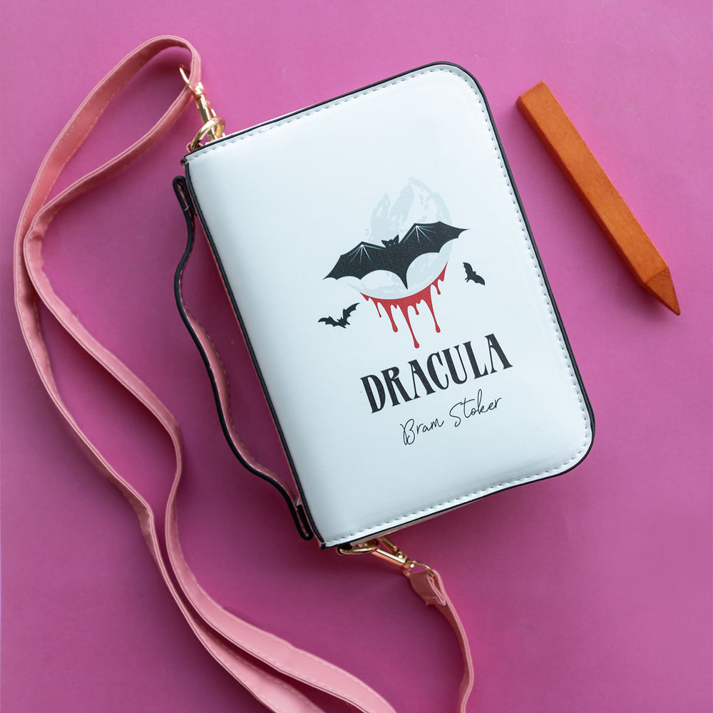 a white purse with black bats and blood on the front, and the words Dracula - Bram Stoker. The purse has a pink strap and is next to a small wooden stake