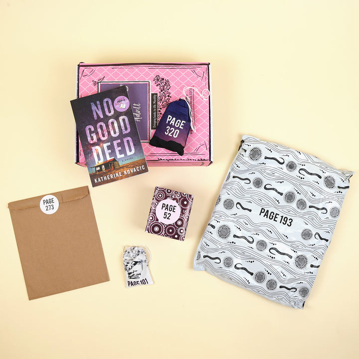 A paperback edition of No Good Deed lays on a pink OUABC box next to a themed wrapped gift labeled with page numbers. In front of the pink OUABC box are four more themed wrapped gifts labeled with page numbers.