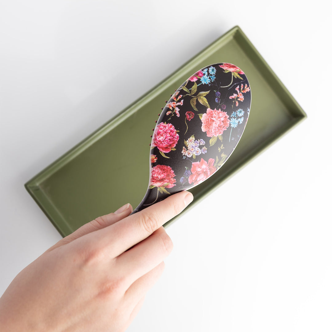 A black hairbrush with floral pattern in shades of pink, orange, blue, white, and green. The hairbrush is held over an open green box.