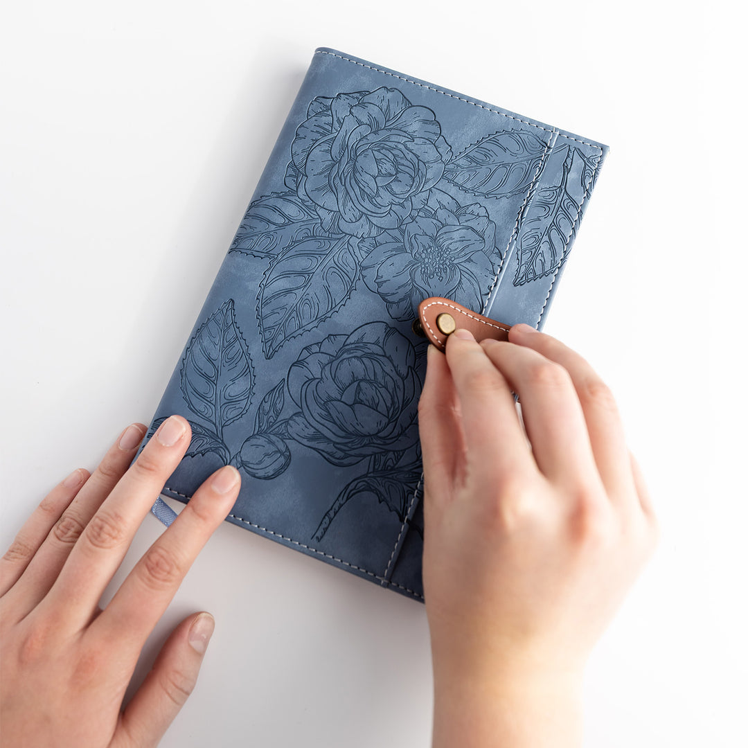 Hands hold a blue sketchbook with a black  floral design and a brown and gold button clasp.