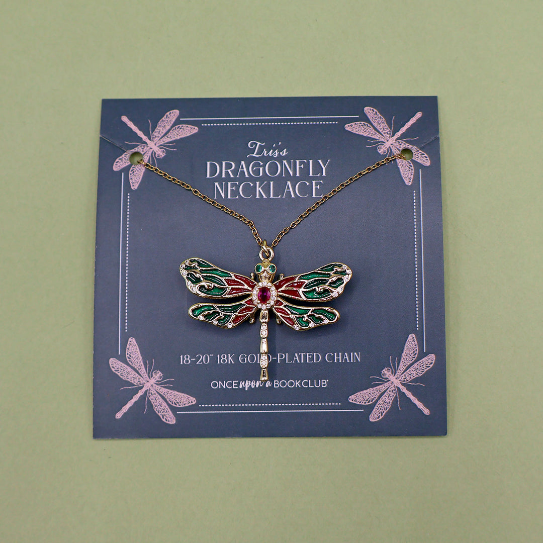 A gold dragonfly necklace attached to a card labeled Iris's Dragonfly Necklace. The dragonfly is made of red and green gems