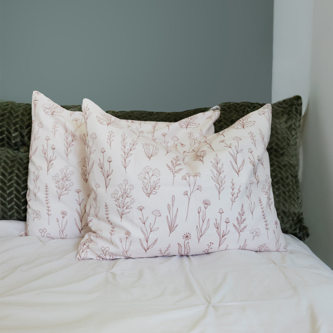Set of two white pillows with a brown flower/plant pattern across each, laying on the bed against a dark green pillow.