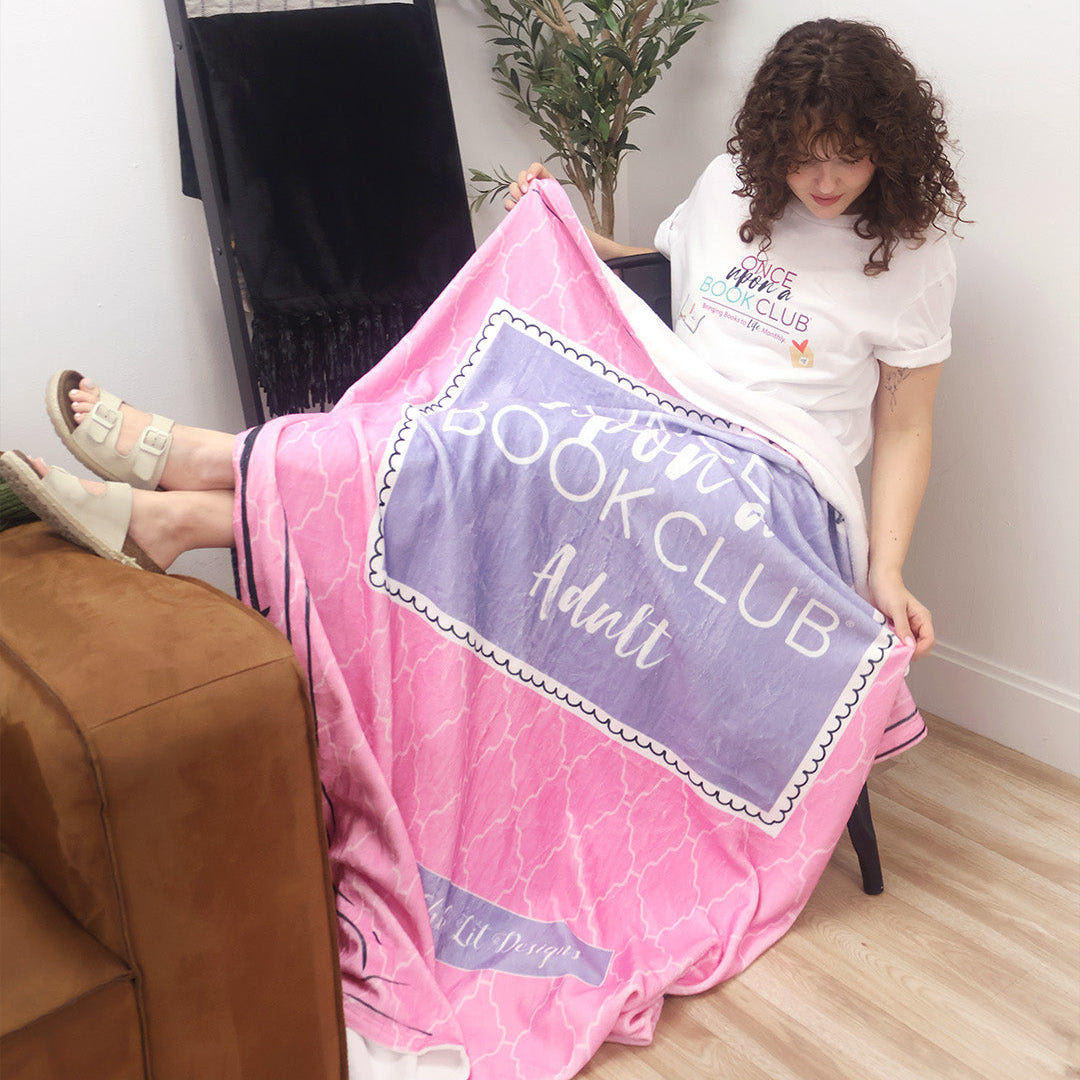 A woman sitting in a chair displays a pink and purple patterned throw blanket inspired by the Once Upon a Book Club Adult box. The scene is set in a bright, modern room with a wooden floor, a black ladder shelf, and a white wall.