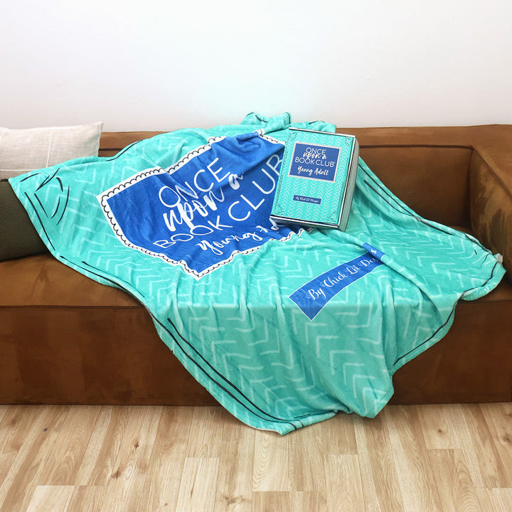 A turquoise and blue patterned throw blanket inspired by the Once Upon a Book Club Young Adult box is draped over a brown leather couch in a living room setting. A matching box sits on the couch with the blanket.