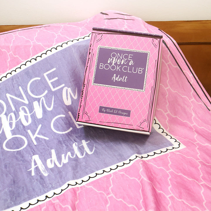 A pink and purple patterned throw blanket inspired by the Once Upon a Book Club Adult box is draped over a brown leather couch in a living room setting. A matching box sits on the couch with the blanket.