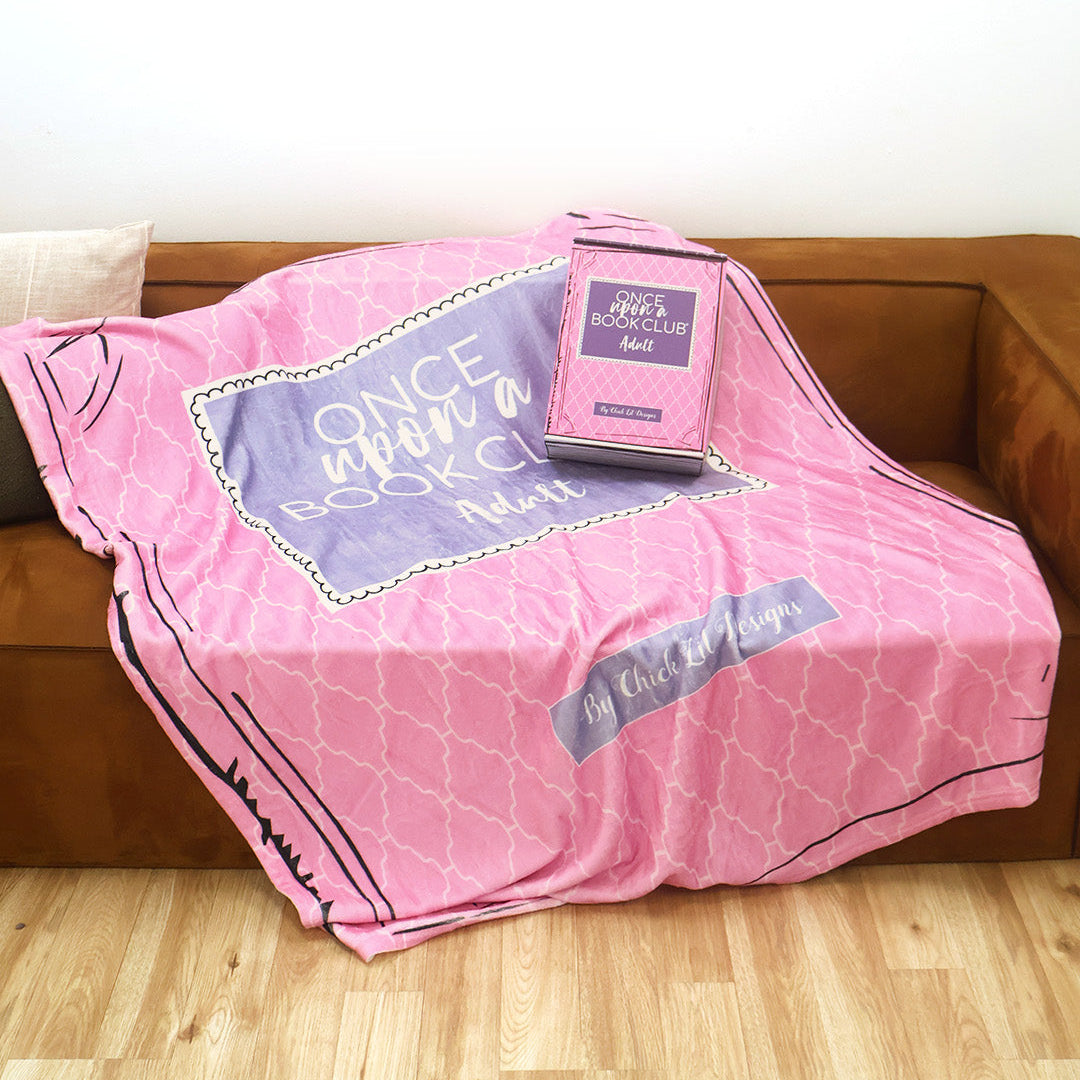 A pink and purple patterned throw blanket inspired by the Once Upon a Book Club Adult box is draped over a brown leather couch in a living room setting. A matching box sits on the couch with the blanket.