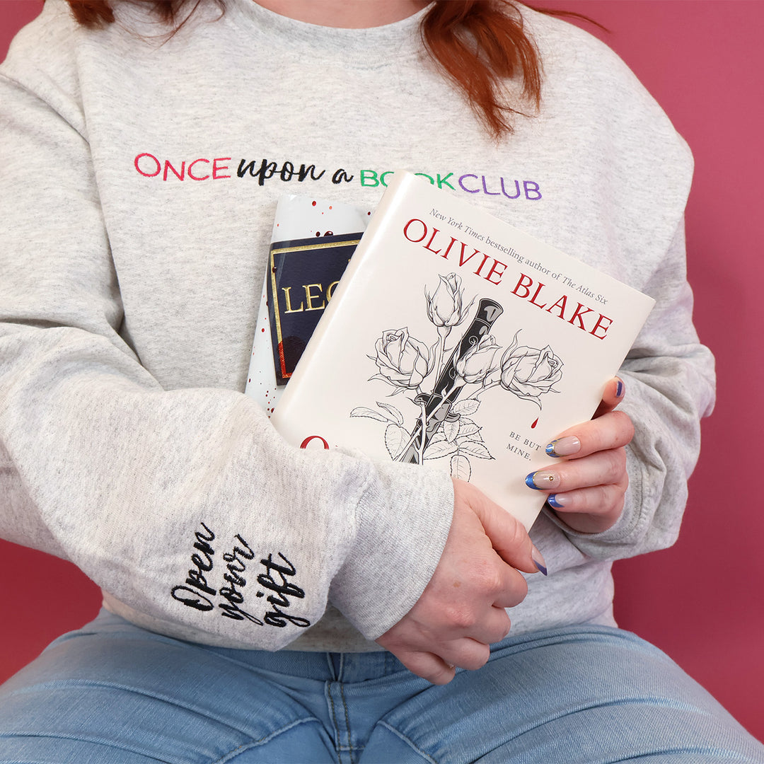 A woman with read hair wearing a light gray sweatshirt with 'Once upon a Book Club' and 'Open your gift' printed on the sleeve, holding a set of books against a solid pink background.