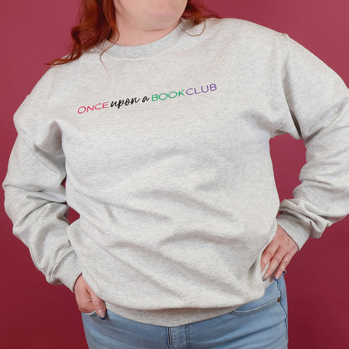 A woman with red hair wearing a light heather-gray sweatshirt with the text 'Once Upon a Book Club' printed across the chest in pink, teal, and purple, paired with light blue jeans against a solid dark pink background.