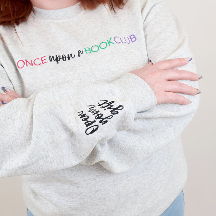 A woman with red hair wearing a light heather gray sweatshirt with colorful text reading 'Once Upon a Book Club' across the chest and black script text reading 'Open Your Gift' on the left sleeve, paired with light blue jeans against a plain light background.