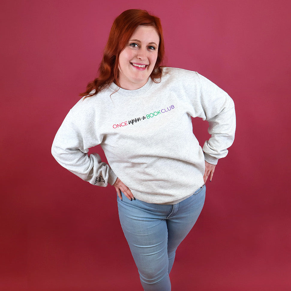 A smiling woman with red hair poses in a heather-gray crewneck sweatshirt featuring the colorful text 'Once Upon a Book Club' on the chest, paired with light blue jeans, set against a solid dark pink studio backdrop.