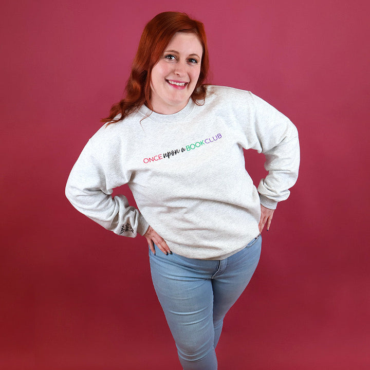 A smiling woman with red hair poses in a heather-gray crewneck sweatshirt featuring the colorful text 'Once Upon a Book Club' on the chest, paired with light blue jeans, set against a solid dark pink studio backdrop.