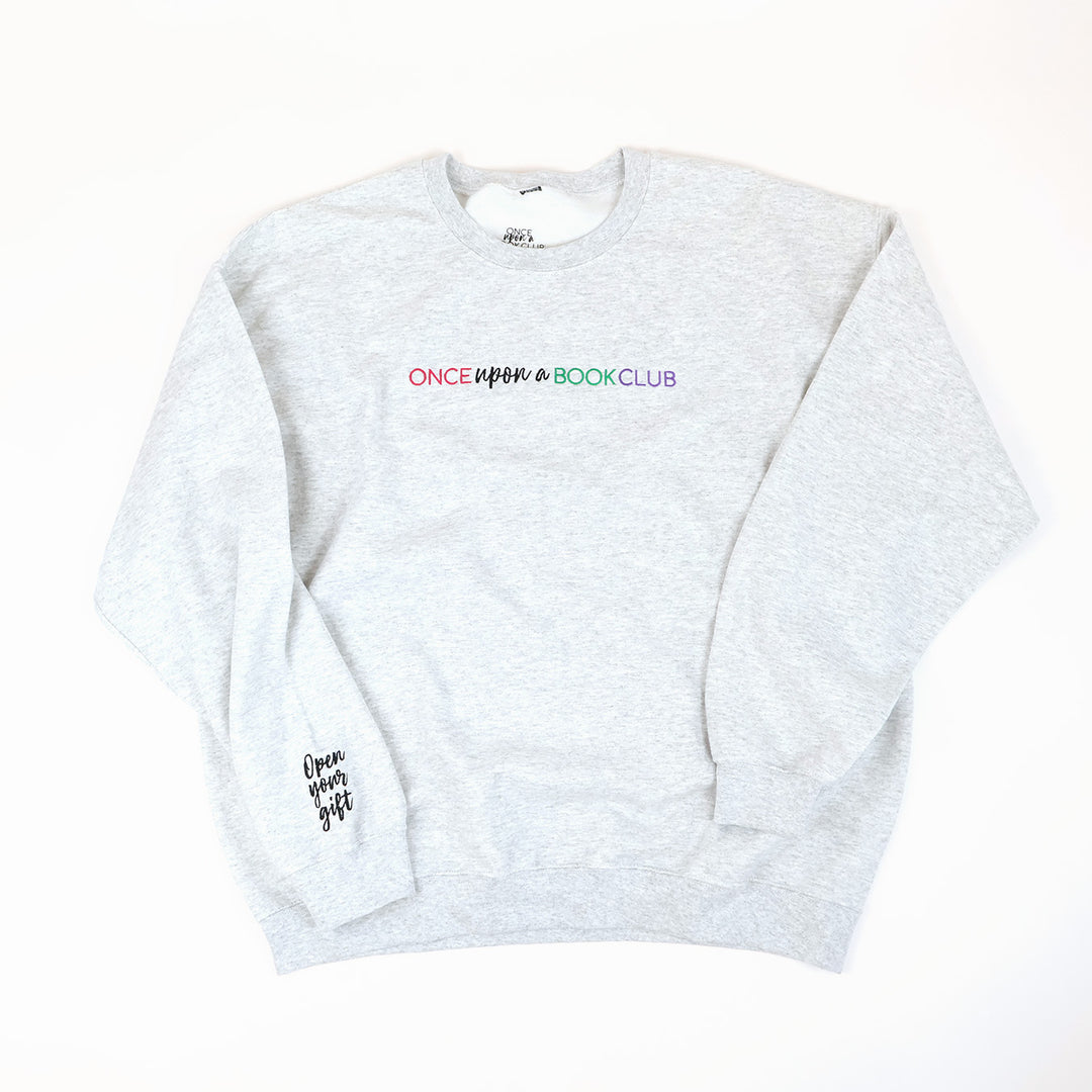 A light heather-gray crewneck sweatshirt with 'Once Upon a Book Club' printed across the chest in a pink, teal, and purple, and 'Open Your Gift' in black script on the left sleeve, laid flat on a white background.
