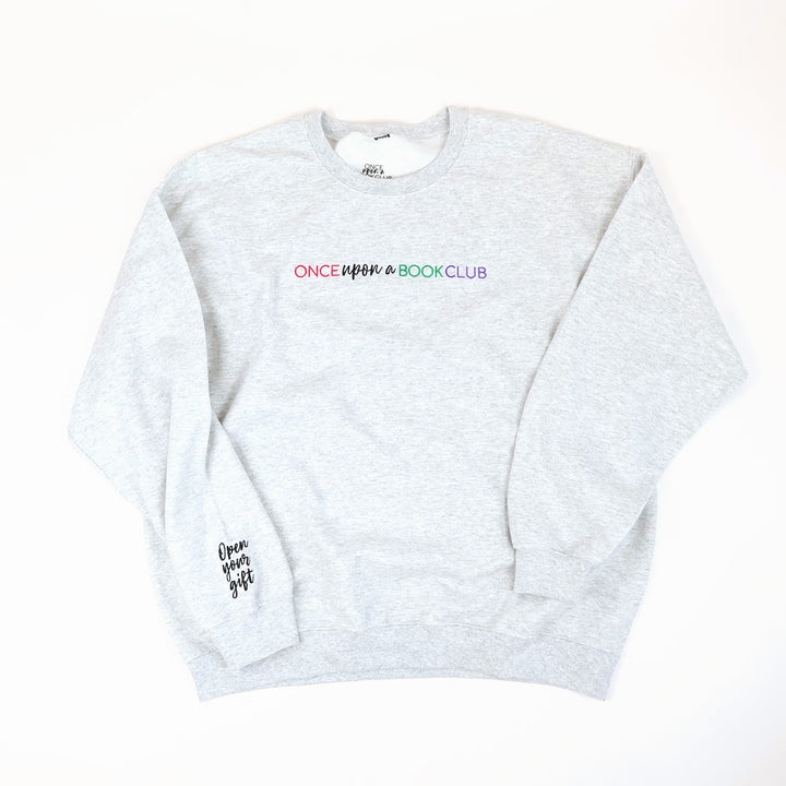 A light heather-gray crewneck sweatshirt with 'Once Upon a Book Club' printed across the chest in a pink, teal, and purple, and 'Open Your Gift' in black script on the left sleeve, laid flat on a white background.