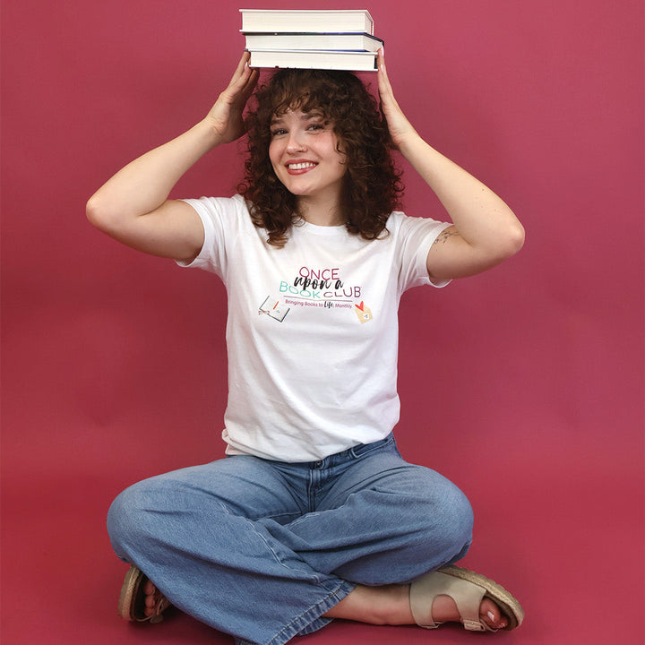 A smiling young woman with curly brown hair sits cross-legged on a pink studio background, balancing a stack of three books on her head. She is wearing a white graphic t-shirt that reads 'Once Upon a Book Club' in pink, teal, and dark purple lettering, blue wide-leg jeans, and beige sandals.