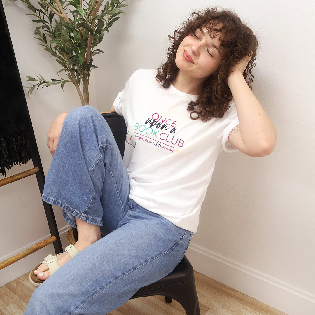 A woman with curly brown hair sits on a black chair wearing a white graphic t-shirt that reads 'Once Upon a Book Club' in pink, teal, and dark purple lettering, paired with light blue wide-leg jeans and beige sandals.