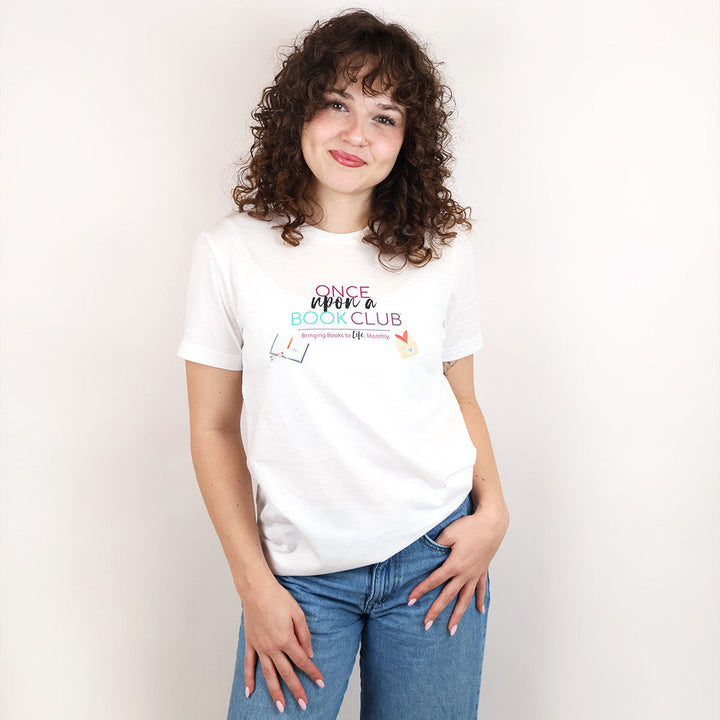 A woman with curly brown hair stands against a plain off-white background wearing a white graphic t-shirt that reads 'Once Upon a Book Club' in pink, teal, and dark purple lettering.