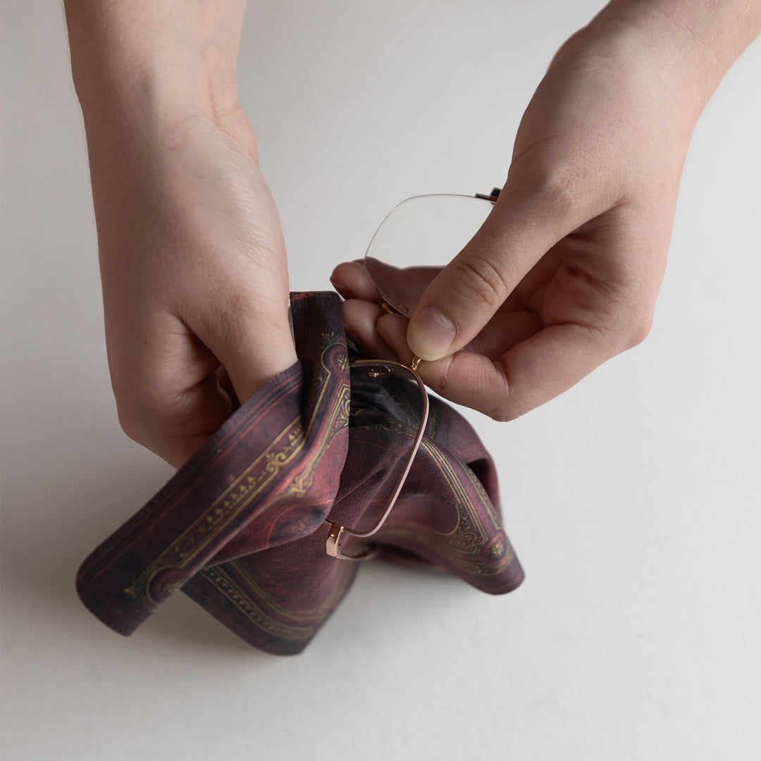 Hands hold a pair of glasses and a screen cleaning cloth that is printed to look like an old book.