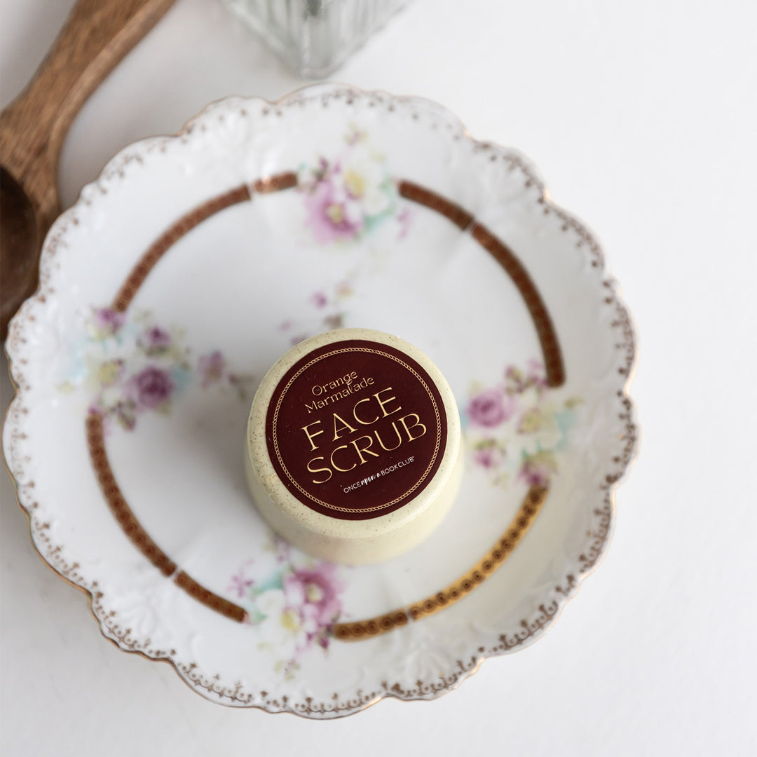 A circular container of Orange Marmalade Face Scrub sits on a white plate.