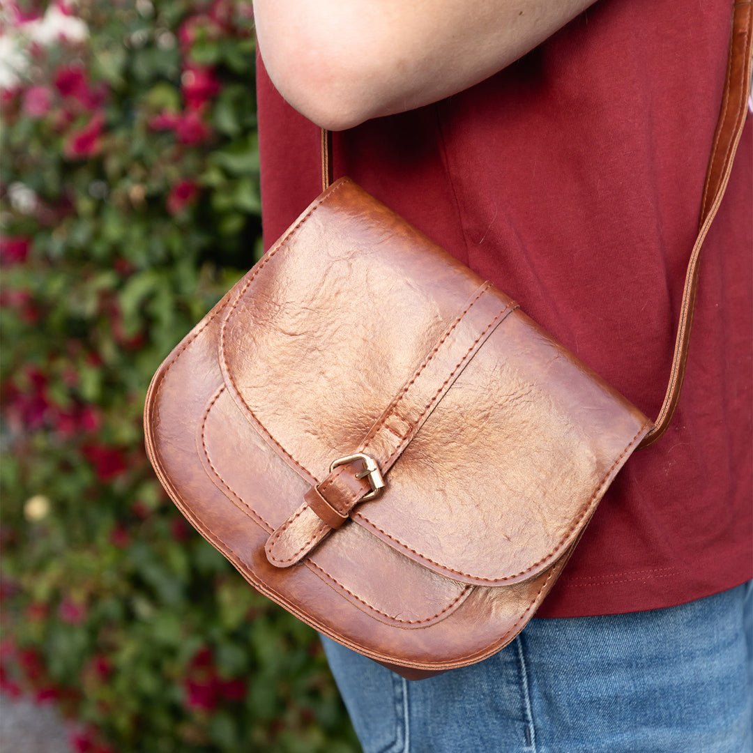 A person with a red shirt and blue jeans wears a brown leather satchel over one shoulder.