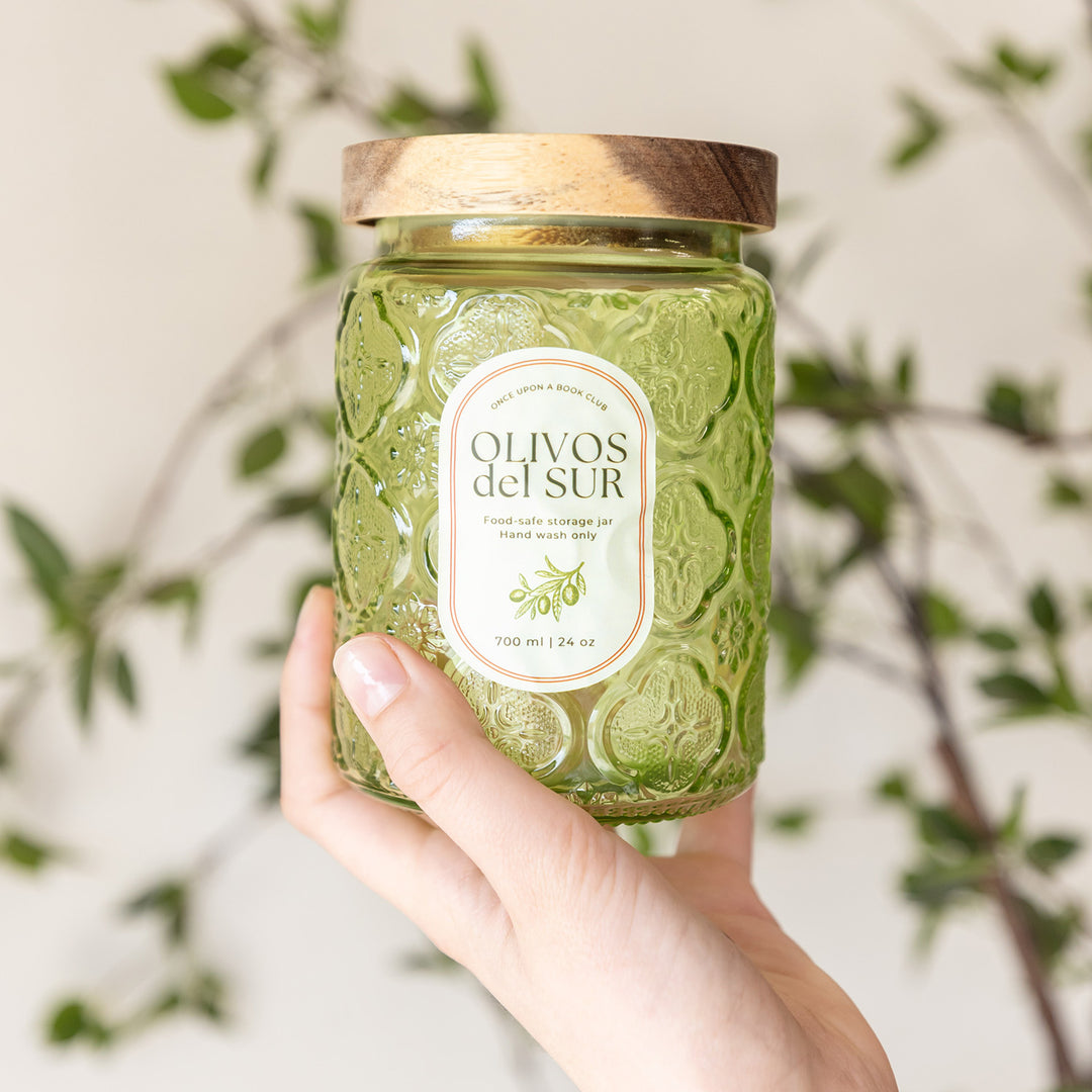 A white hand holds up a green transparent Olive jar with a wooden lid. 