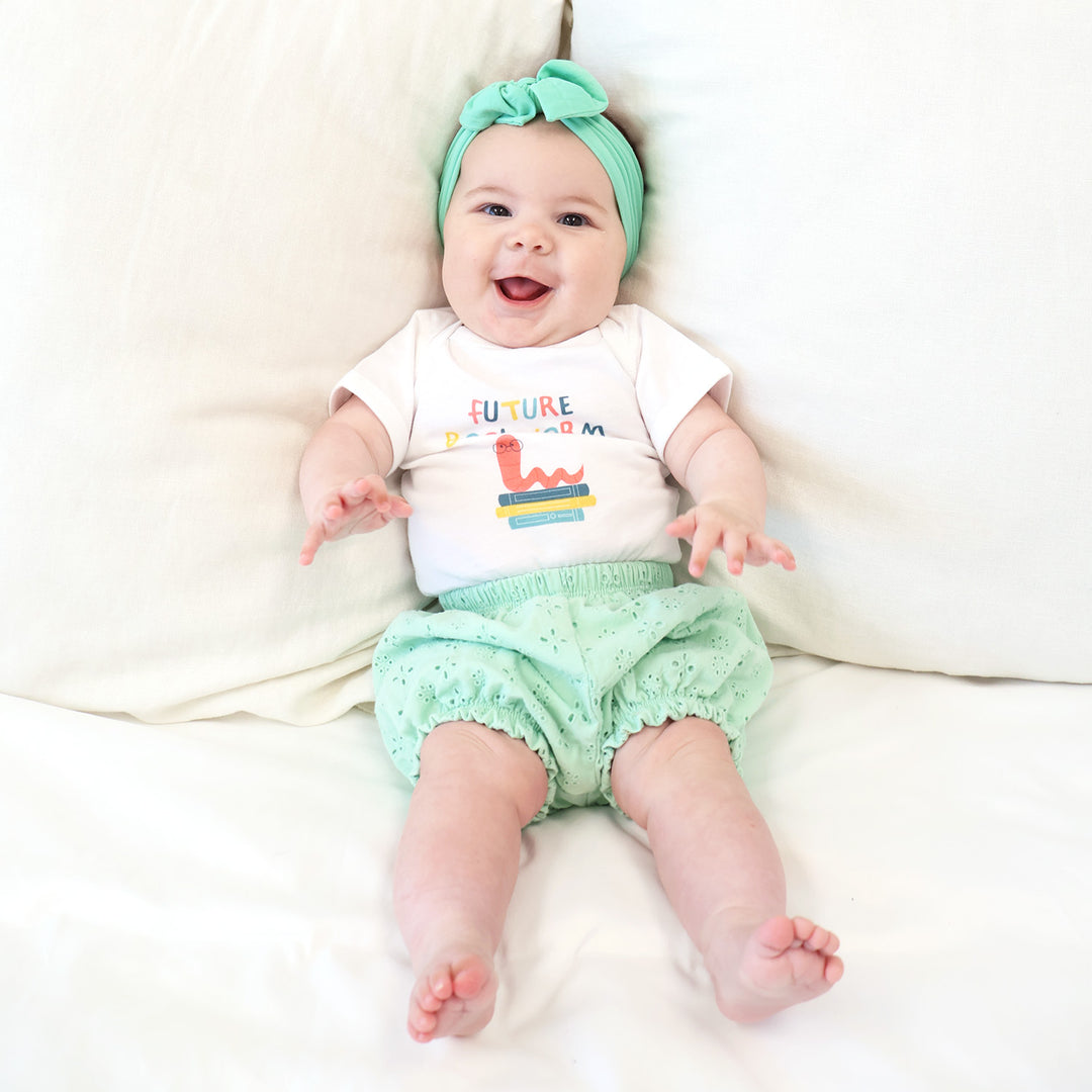 Baby wearing a 'Future Bookworm' shirt and green headband and floral shorts on a white blanket