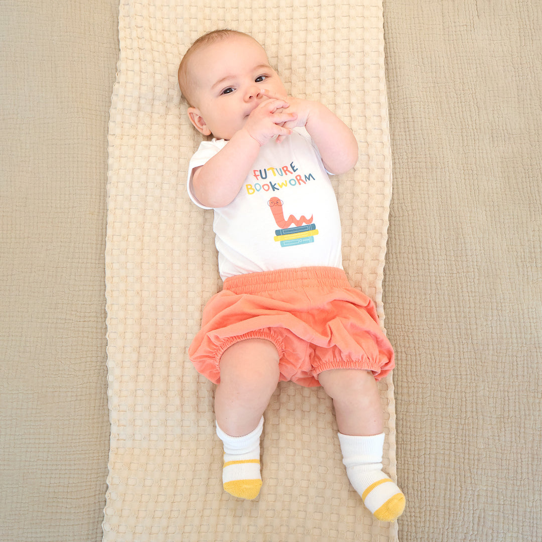 Baby lying on a textured blanket wearing a 'Future Bookworm' shirt and orange shorts.