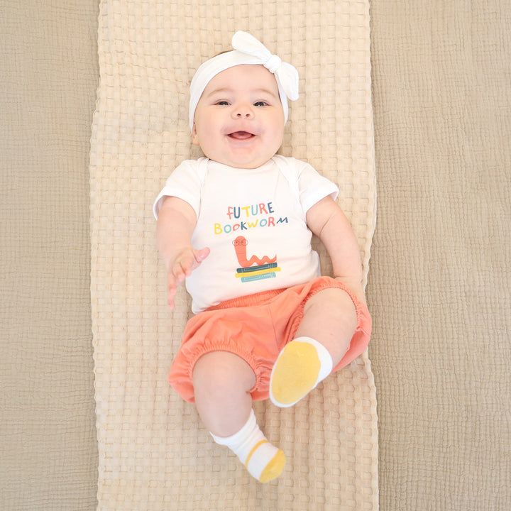 Baby wearing a 'Future Bookworm' shirt and orange shorts on a beige blanket