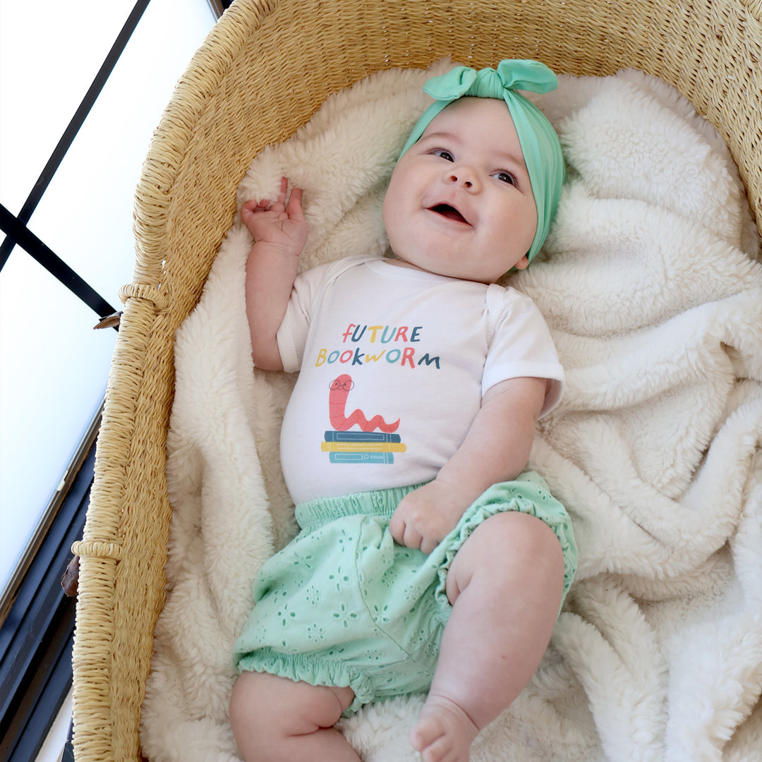 Baby in a bassinet wearing a 'Future Bookworm' shirt and green headband.
