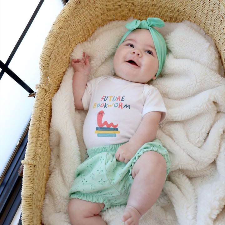 Baby in a bassinet wearing a 'Future Bookworm' shirt and green headband.