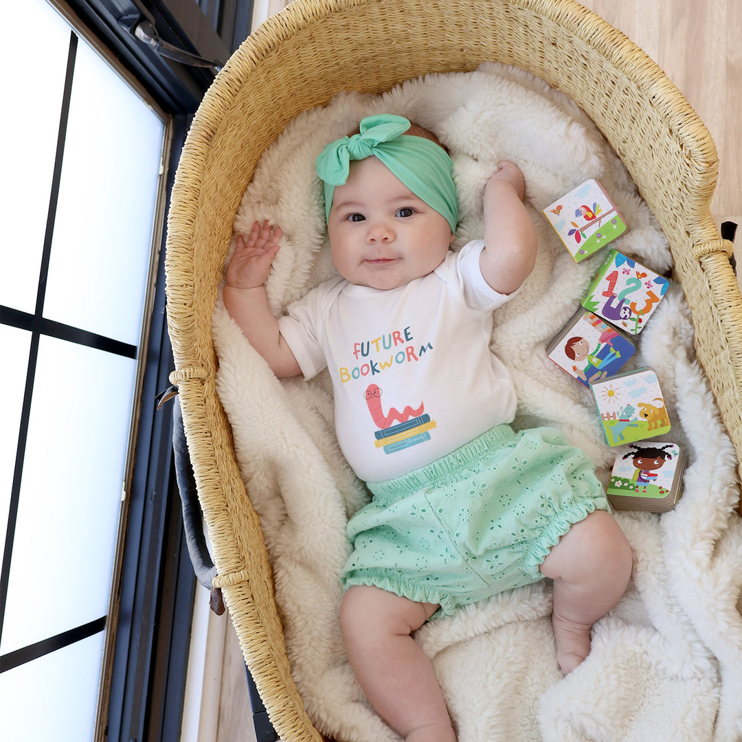 Baby in a bassinet wearing a 'Future Bookworm' shirt and green headband, surrounded by children's books.
