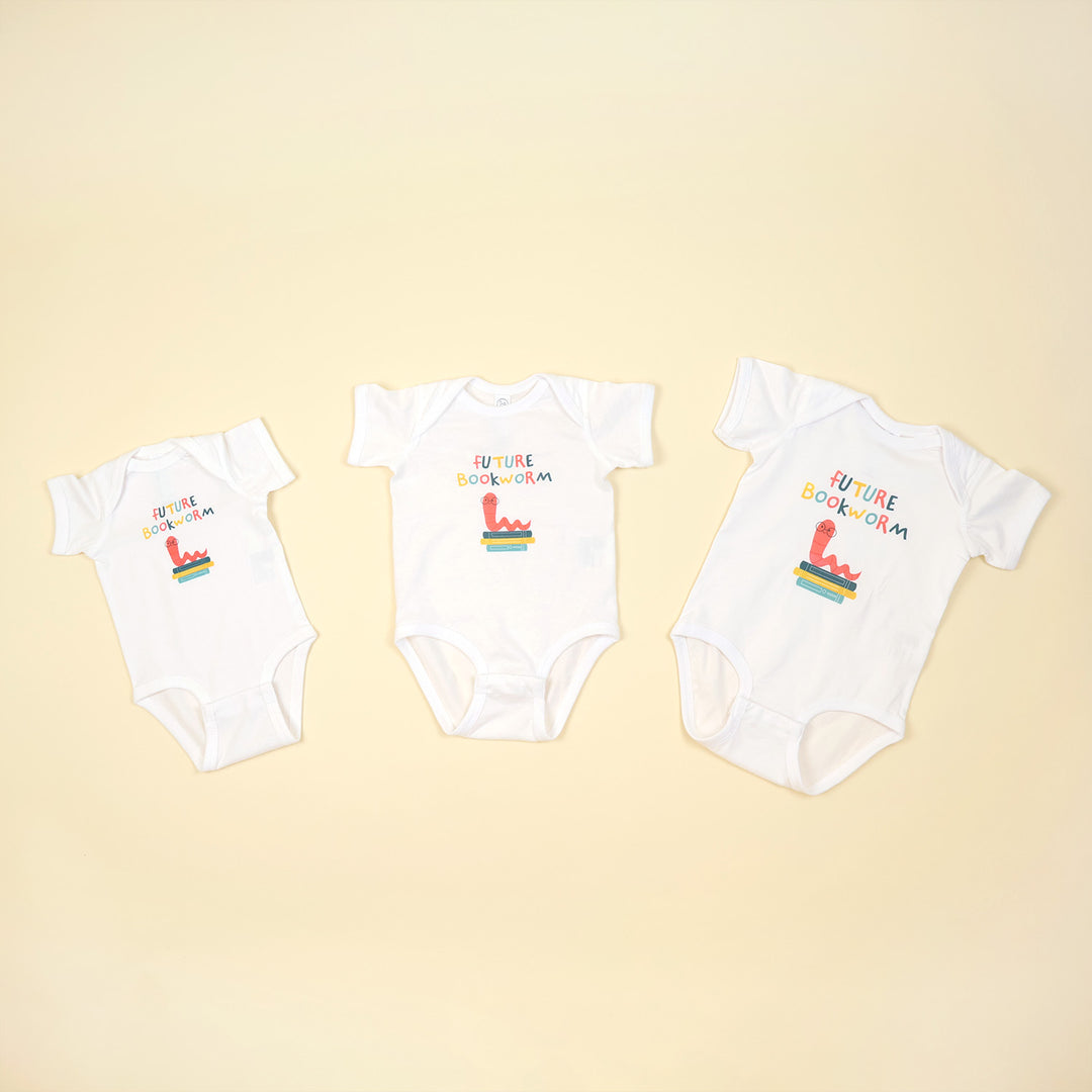 Three white baby onesies with colorful text and graphics on a beige background