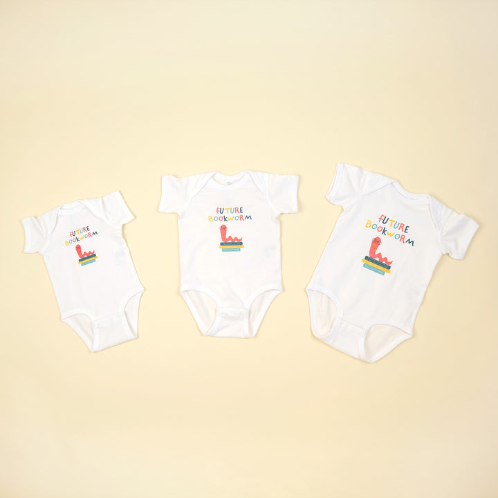 Three white baby onesies with colorful text and graphics on a beige background