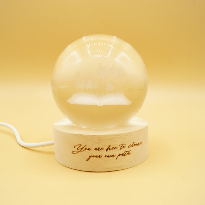 a glass globe light featuring an open book with trees engraved inside the orb. There is a wooden base burned with the quote 'You are free to choose your own path'