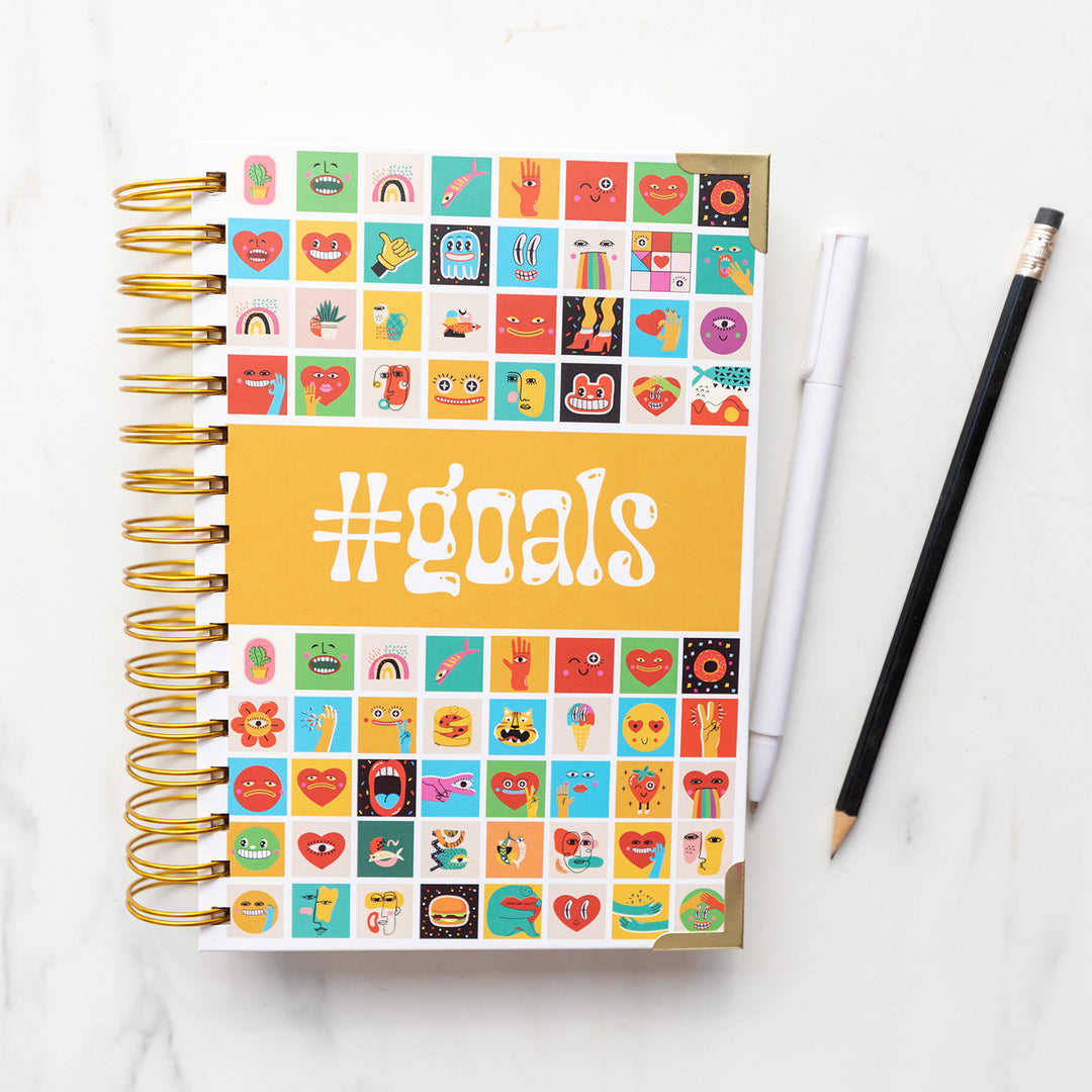 A #goals lined notebook perfect for writing down all your hopes and dreams! The cover features a bunch of fun and quirky icon faces. A black pencil and white pen sit next to the ringed notebook.
