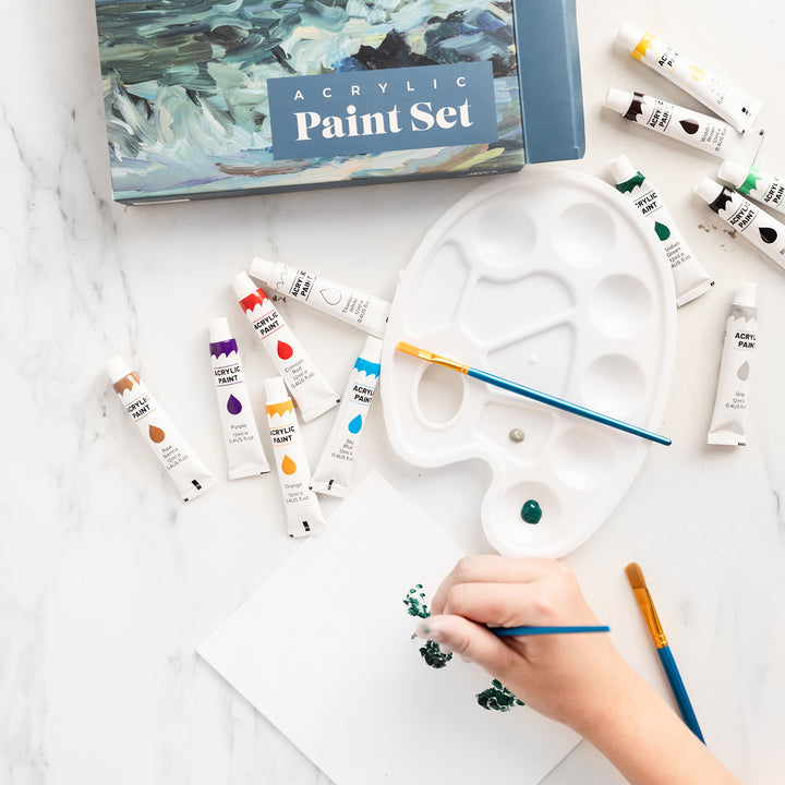 a white hand holding a brush paints on a white canvas. Acrylic paint tubes are scattered over the marble surface along with a white painter's palette and two more brushes